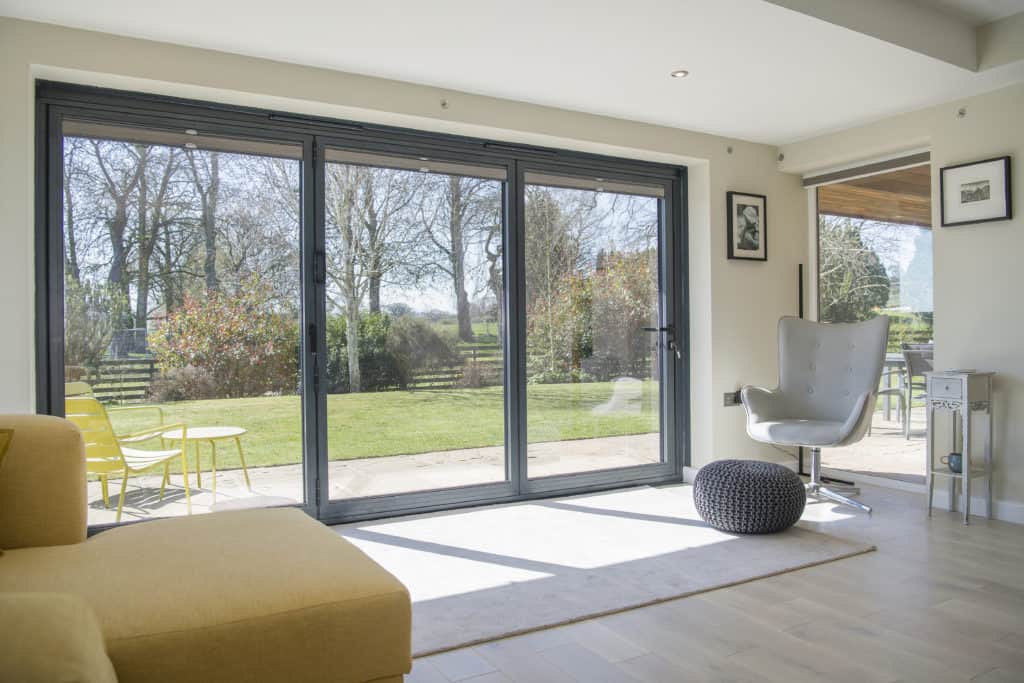 A modern living room with large bifold doors opening to a sunny garden. The room features a yellow sofa, a gray chair, a round ottoman, and light wood floors. Outdoor seating and trees are visible outside.