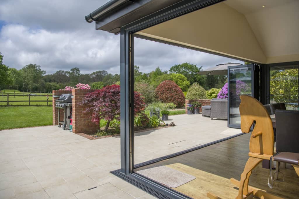 View from inside a house through expansive bifold doors to a patio with outdoor seating, a barbecue grill, and a garden filled with colorful shrubs and green trees under a partly cloudy sky.
