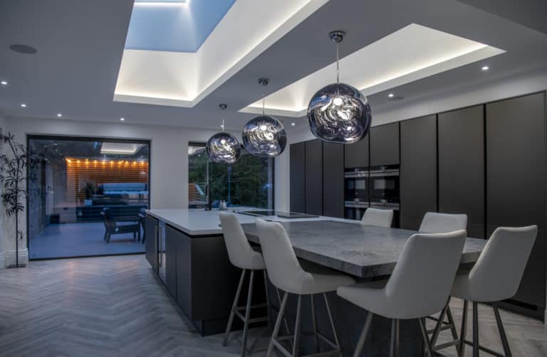 Modern kitchen with a large island, white chairs, dark cabinets, pendant lights, skylights, and bifold doors leading to an outdoor seating area. The space is brightly lit with both natural and artificial lighting.