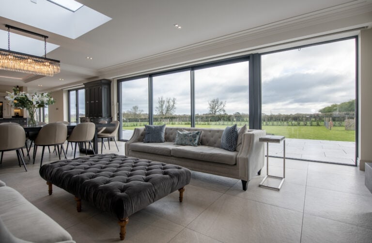 Modern open-plan living room with large floor-to-ceiling windows and sliding doors, light grey sofas, a dark ottoman, dining area with white chairs, and a view of a green lawn and trees outside under a cloudy sky.