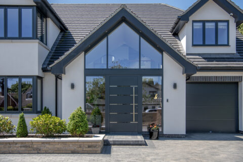 Modern house exterior with bespoke glass solutions, featuring a large glass front door, tall peaked roof, grey trim, symmetrical windows, a driveway, small garden beds with shrubs, and a single-car garage on the right.