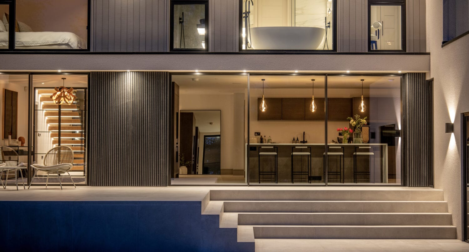 Modern home exterior at night with bespoke glass solutions like large sliding doors, an illuminated kitchen and dining area, pendant lights over a kitchen island, and a freestanding bathtub visible through an upstairs window. Wide steps lead up to the patio.