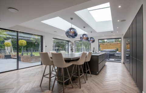 Modern kitchen with large skylights, an island with four high chairs, pendant lights, and floor-to-ceiling bespoke glass solutions including sliding doors opening to a patio and garden, allowing natural light to fill the space.