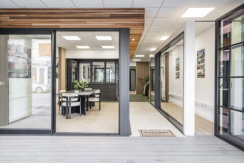 Modern office space with large glass doors and bespoke glass solutions, a round meeting table with chairs, indoor plants, wood and metal accents, and bright overhead lighting. A “Welcome” mat is placed at the entrance.