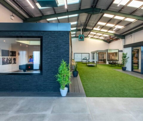 A modern indoor office space with artificial grass, glass-walled meeting rooms, a ping pong table, potted plants, and large windows with sliding doors letting in natural light.