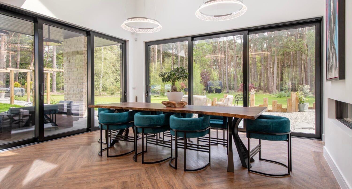 A modern dining room with a large wooden table, teal velvet chairs, and floor-to-ceiling bifold doors offering views of a lush garden and forest. The space features wood flooring and two circular pendant lights overhead.