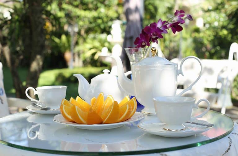 A glass table set outdoors with a white teapot, two teacups and saucers, a plate of orange slices, and a vase with purple orchids. Green garden foliage is blurred in the background.