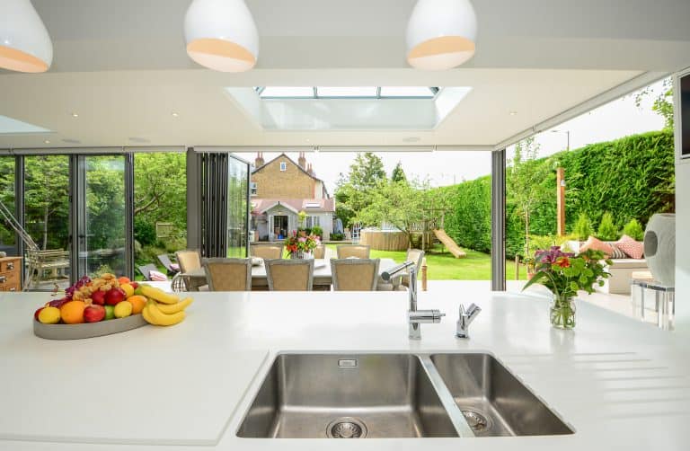 Modern kitchen with a white island, double sink, fruit bowl, and vase of flowers. Large glass doors open to a patio with outdoor dining, green garden, and childrens play area. Natural light fills the space.