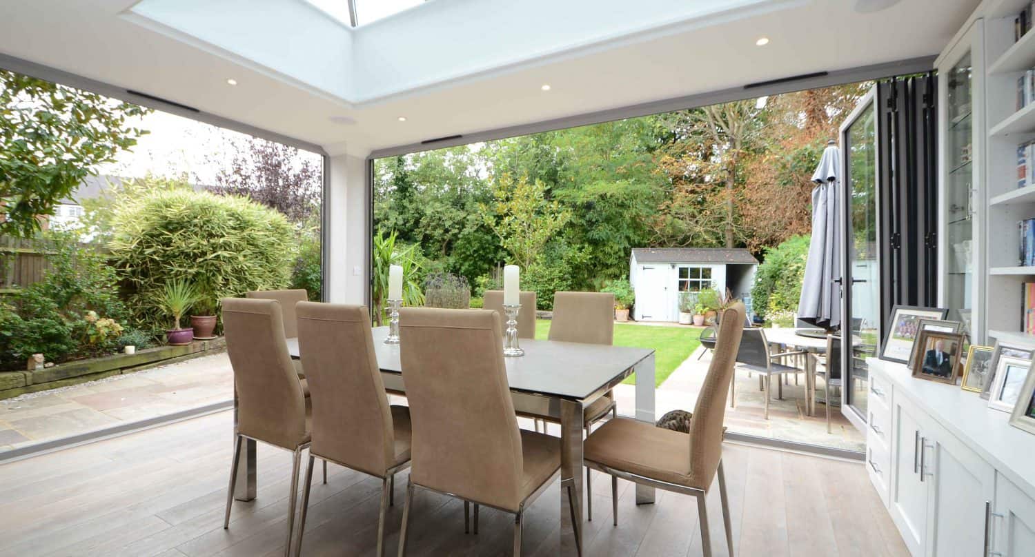 Modern dining room with beige chairs around a dining table, large skylight, and glass doors open to a lush green garden with a patio, tall plants, and a small white shed in the background.