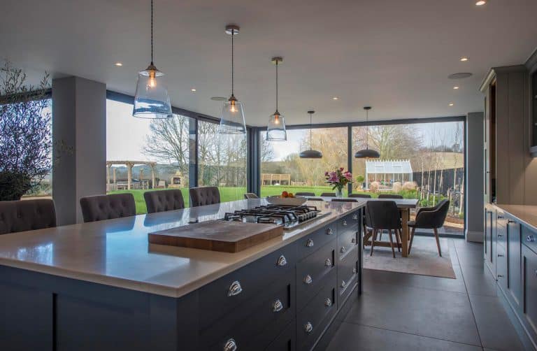 Modern kitchen with a large island, pendant lights, gas cooktop, and views of a garden through floor-to-ceiling windows. Dining table with chairs and fresh flowers is visible in the background.