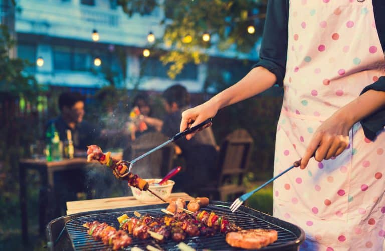 A person wearing a pink polka dot apron grills skewers and meat at an outdoor barbecue, with friends sitting and talking in the background under string lights.
