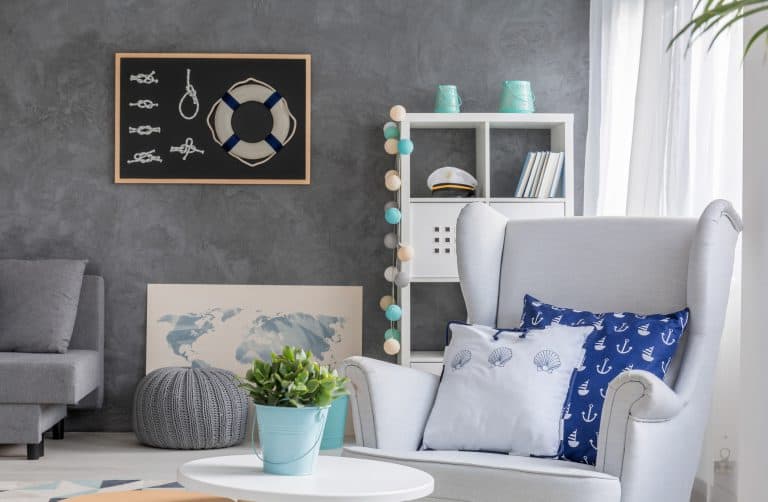 A cozy nautical-themed living room with gray walls, a white armchair, blue and white pillows, a potted plant, a chalkboard with knots, and a shelving unit with decor including a captain’s hat and string lights.