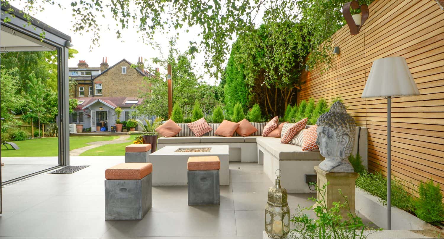 A modern outdoor patio features a white sofa, orange cushions, concrete stools, fire pit, Buddha statue, floor lamp, and wooden slat privacy fence. Bespoke glass solutions showcase lush greenery and offer a view of the house in the background.
