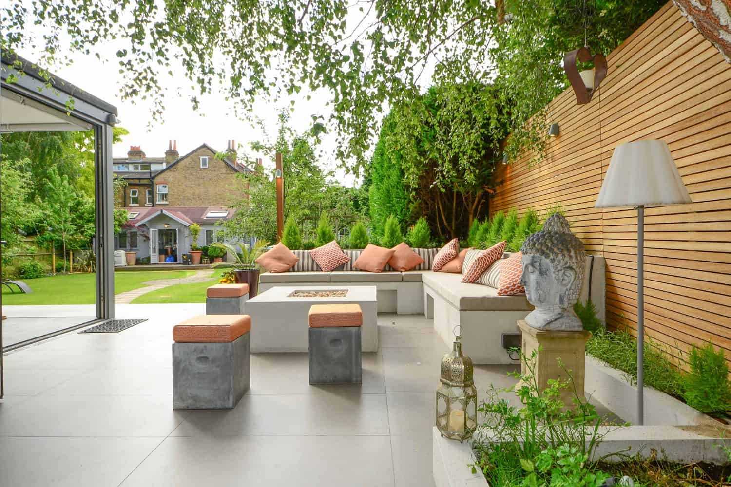 Modern outdoor patio with a white L-shaped sofa, orange cushions, concrete stools, a Buddha head statue, lantern, floor lamp, and wooden privacy fence. Lush greenery and houses are visible in the background.