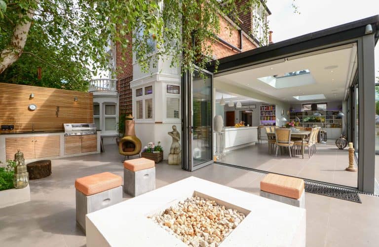 A modern outdoor patio with a fire pit, seating, a barbecue grill, and a wood wall. Sliding glass doors open to a bright kitchen and dining area, with large trees providing shade.