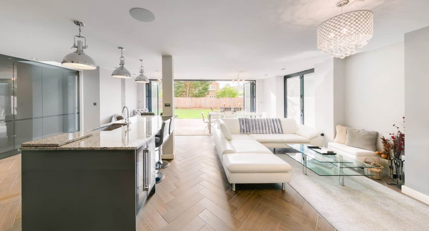 Modern open-plan kitchen and living room with a white sectional sofa, glass coffee table, large windows, pendant lights, and sliding doors leading to a backyard with a lawn and trees.