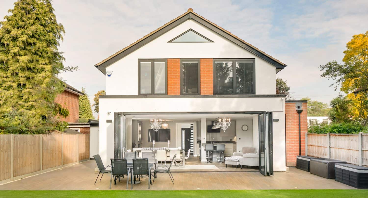 A modern two-story house with large windows and folding glass doors opening to a patio with outdoor dining furniture, surrounded by trees and a fenced yard.