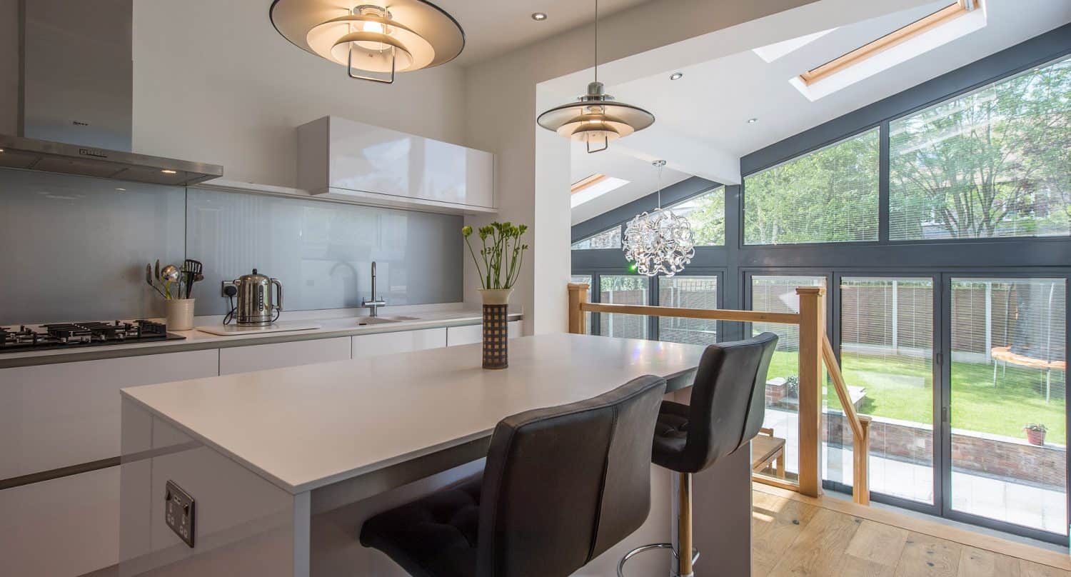 Modern kitchen with a white island, two black chairs, pendant lights, and large windows letting in natural light. The view outside shows a green garden and a wooden fence.