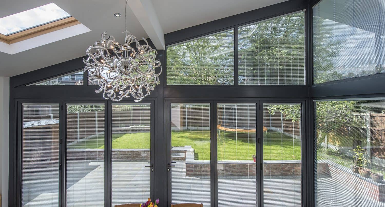 Modern sunroom with large glass sliding doors and windows looking out onto a green lawn and patio; a decorative chandelier hangs from the ceiling above a wooden table with a vase of tulips.