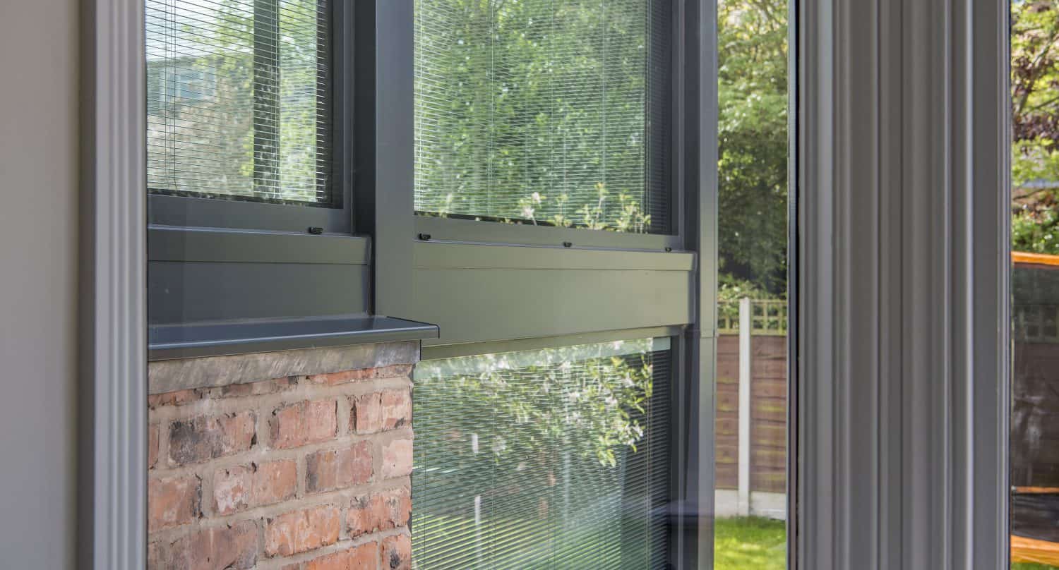 View from inside a building showing a large window with green metal frames, a section of exposed brick wall, and bifold doors opening to a garden with trees and grass visible outside.