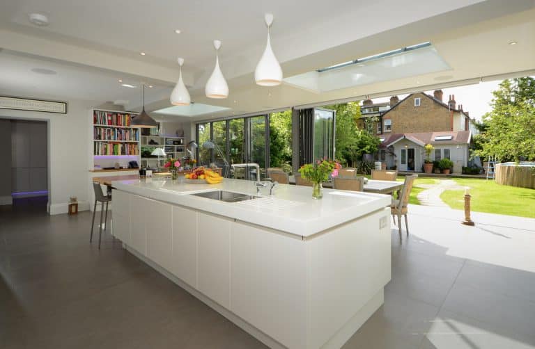 Modern, bright kitchen with a large white island, hanging pendant lights, bookshelves, and glass doors opening to a green garden with a path leading to another house. Flowers and fruit are on the countertop.