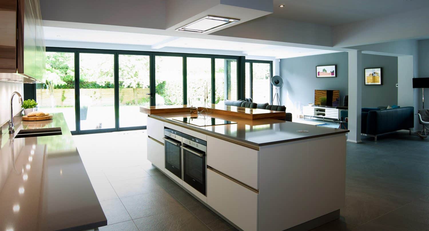 Modern open-plan kitchen with a large white island, built-in ovens, and sleek counters. Floor-to-ceiling glass doors open to a patio, allowing natural light to fill the spacious living and dining area.