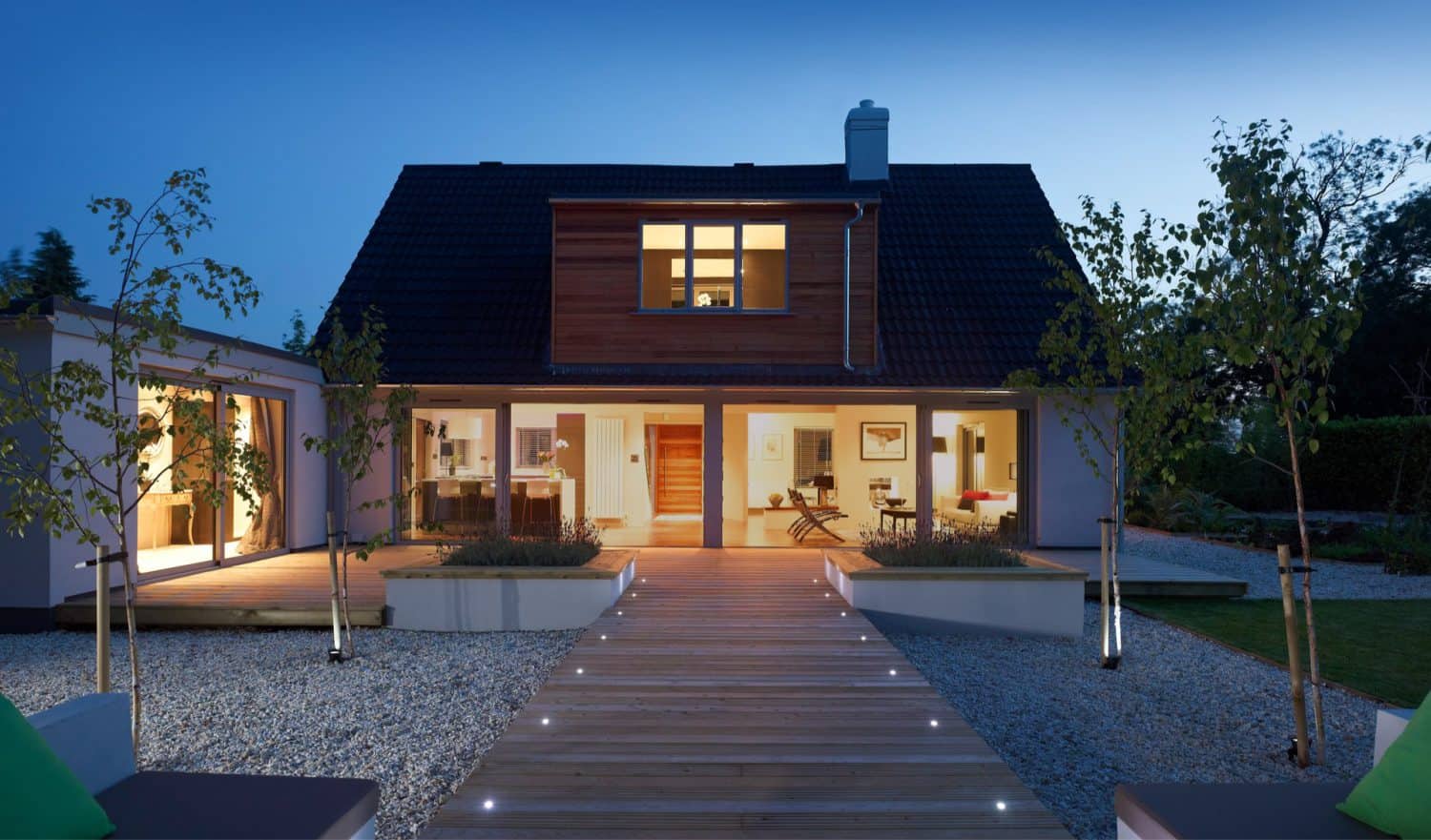 Modern house at dusk with large windows glowing warmly, wooden deck pathway with lights leading to the entrance, landscaped with young trees and gravel.