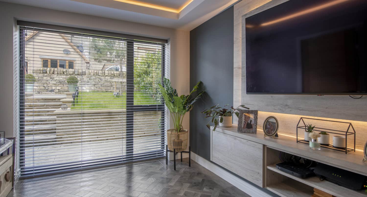 Modern living room with bespoke glass solutions, large window blinds, herringbone wood floor, wall-mounted TV, plants, and shelves with framed photos and decor; view of a garden and stone wall outside.