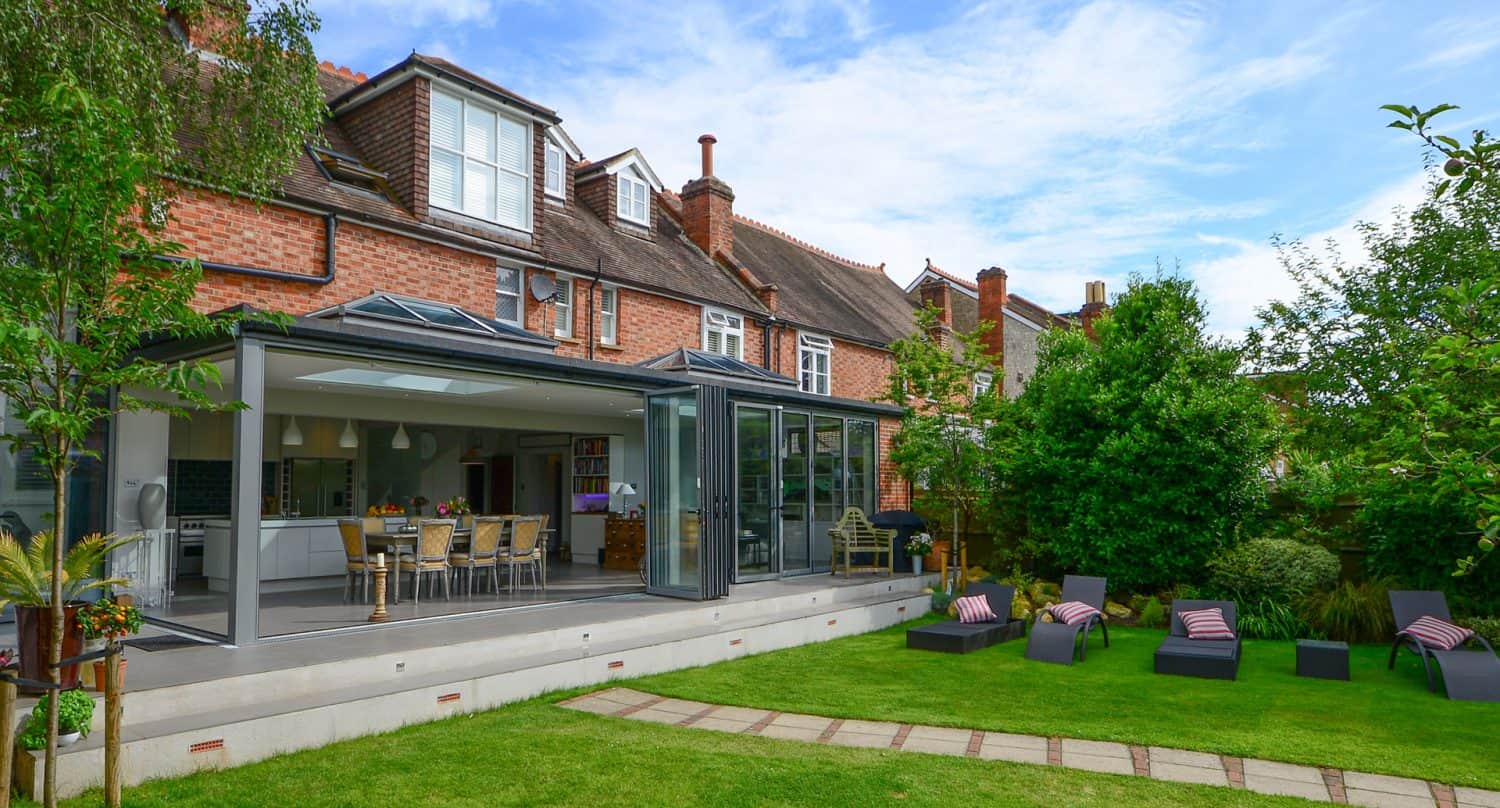 A modern house with a large glass extension opens to a patio with a dining table and chairs. The well-kept backyard has green grass, lounge chairs with striped cushions, and surrounding trees and shrubs.