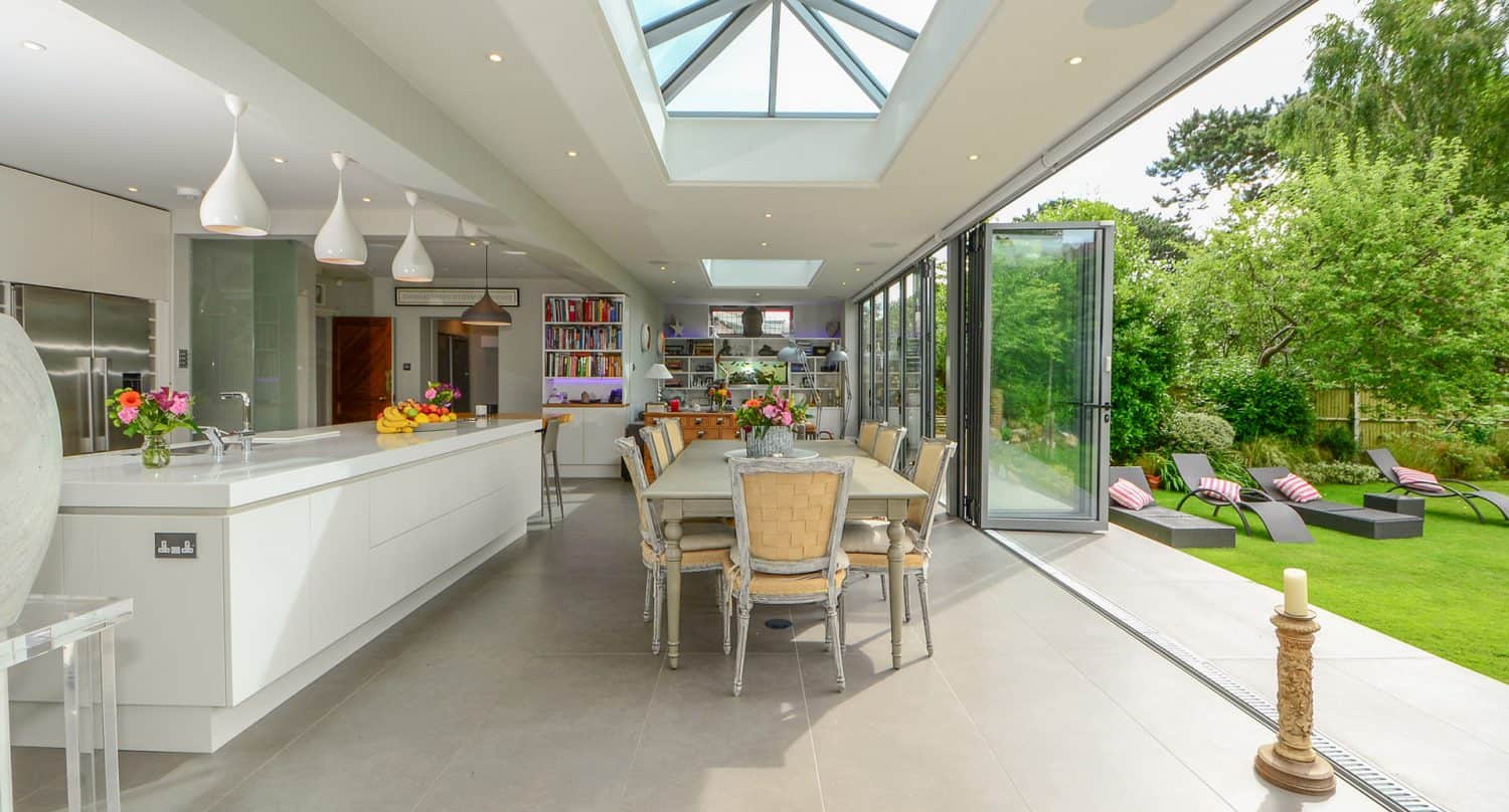 Modern open-plan kitchen and dining area with skylight ceiling and large glass doors opening to a green garden with lounge chairs; elegant decor and fresh flowers on tables.