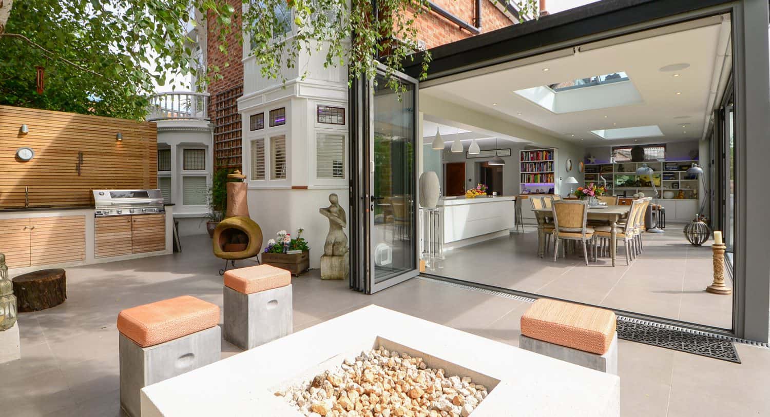 Modern open-plan kitchen and dining area with large glass doors opening to a stylish patio featuring a fire pit, outdoor seating, built-in grill, and greenery for a seamless indoor-outdoor living space.