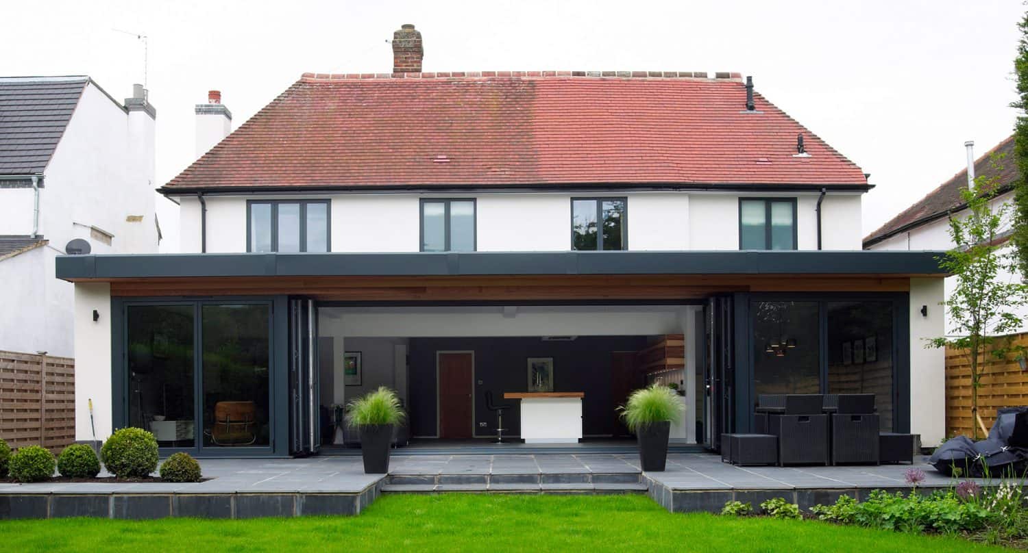 A modern house with large sliding glass doors, a red tile roof, and a spacious patio area. The patio features potted plants and outdoor seating, overlooking a green lawn.