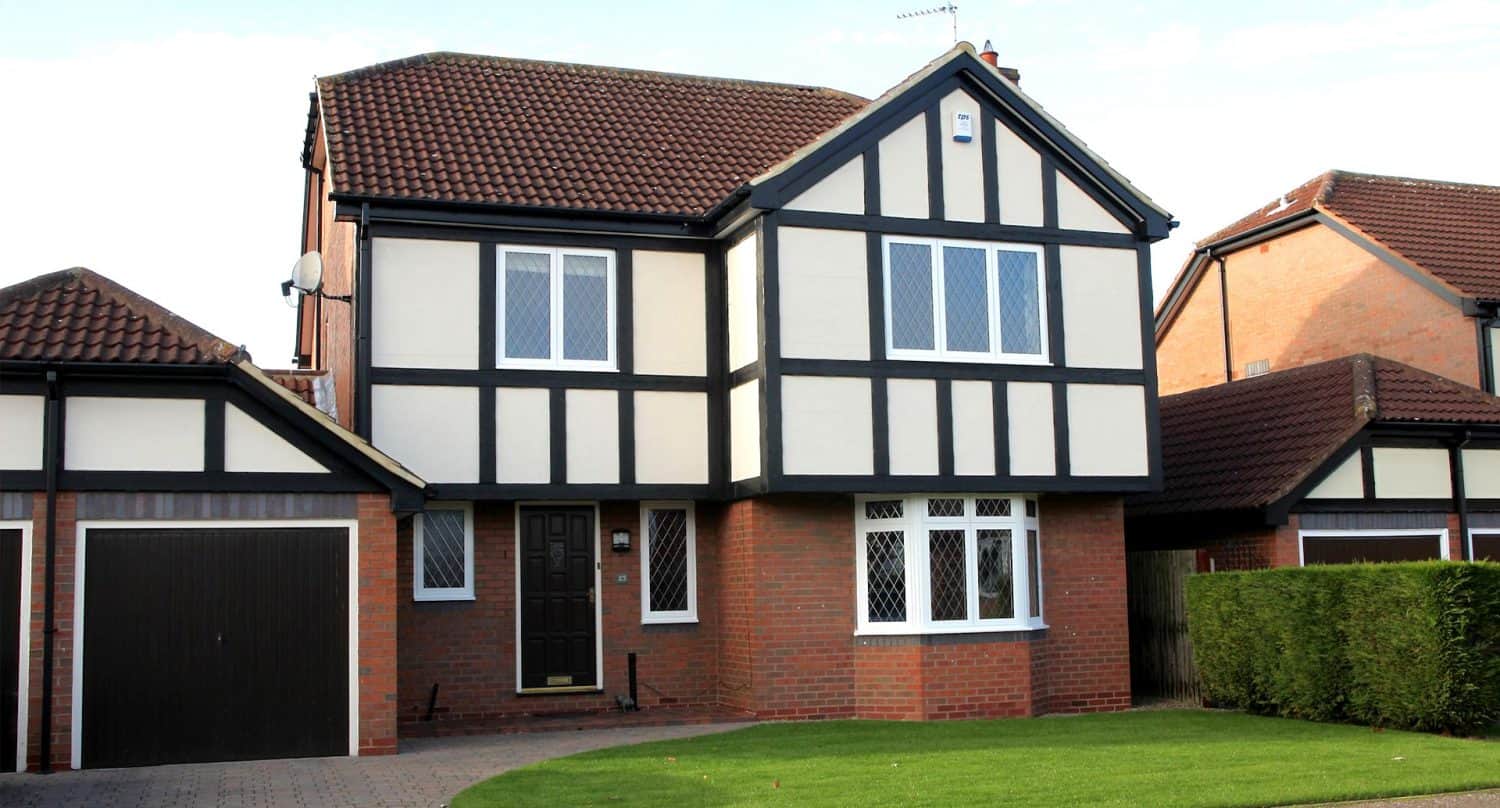 A two-story Tudor-style house with a brown tiled roof, black and white exterior timber detailing, brick lower level, a dark front door, large windows, attached garage, and a neatly trimmed front lawn.