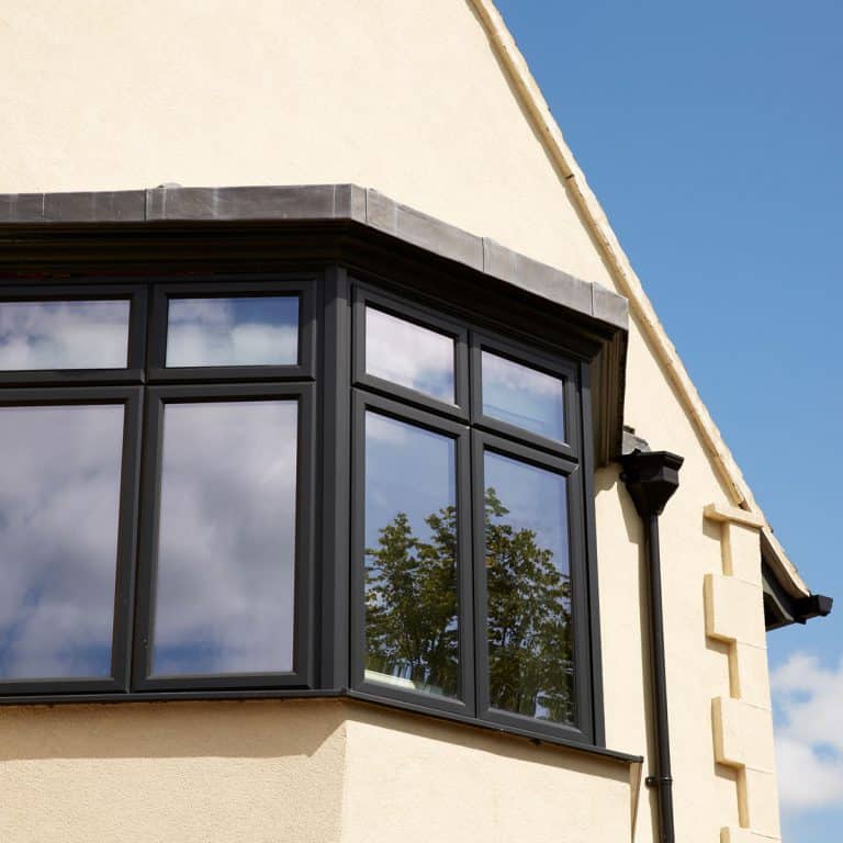 A close-up of a cream-colored house’s black-framed bay window, reflecting trees and sky in the glass, under a clear blue sky. A black drainpipe runs down the house’s corner.