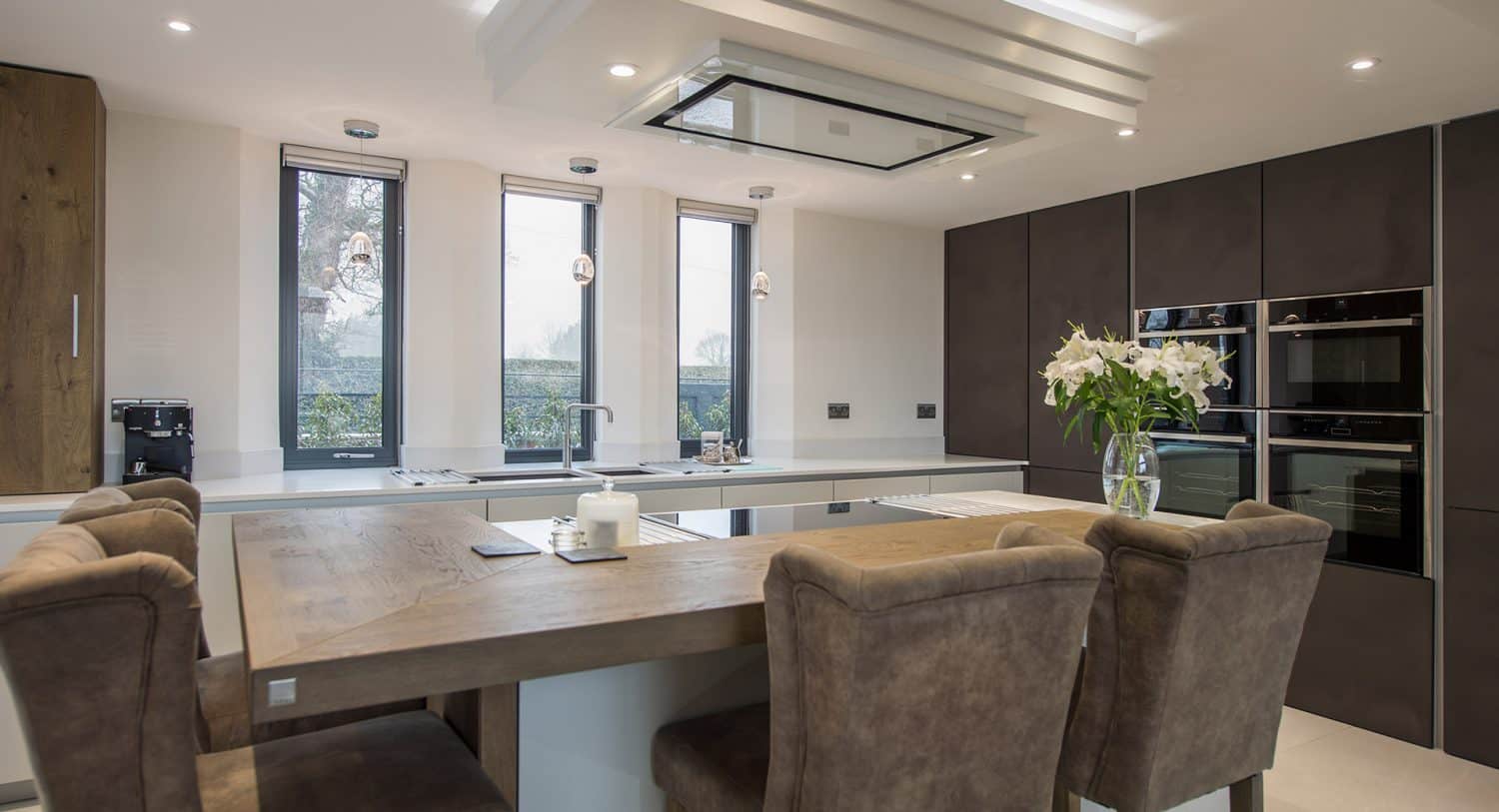 Modern kitchen with a wooden dining table, four upholstered chairs, built-in ovens, dark cabinets, a kitchen island with a sink, large windows, and a vase of white flowers.