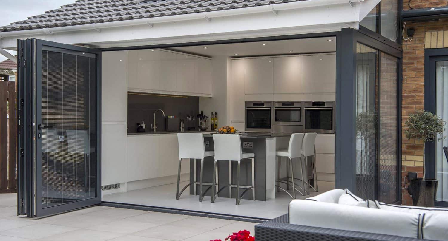 Modern kitchen with white cabinets and island, four white barstools, and built-in ovens. Glass folding doors open to a patio with outdoor seating, creating a seamless indoor-outdoor living space.