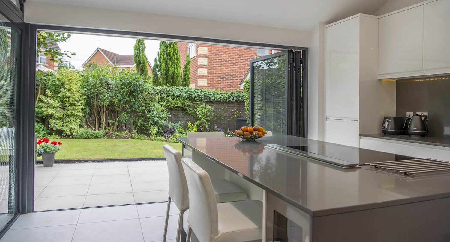 A modern kitchen with a glossy island and stools opens to a lush green garden through large folding glass doors, letting in natural light. A bowl of fruit sits on the island.