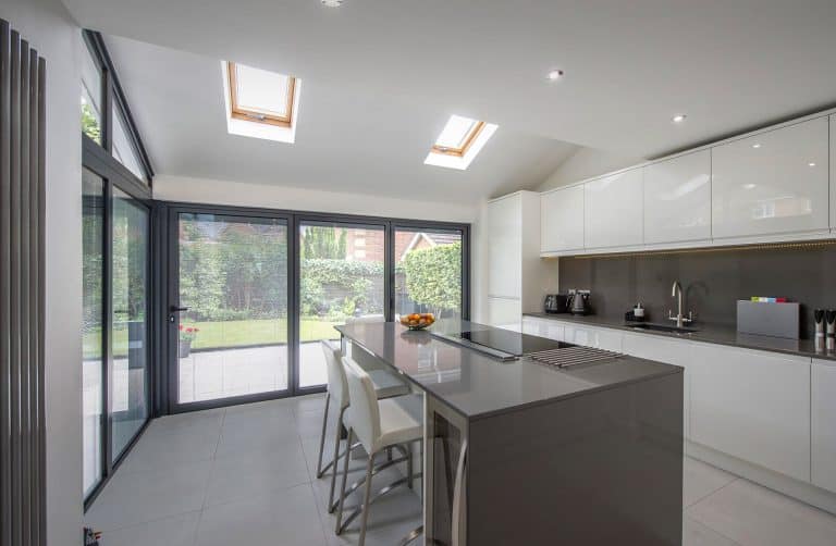 Modern kitchen with white cabinets, a gray island with built-in cooktop, four white bar stools, skylights, large glass doors, and a view of a green backyard. A bowl of oranges sits on the island.
