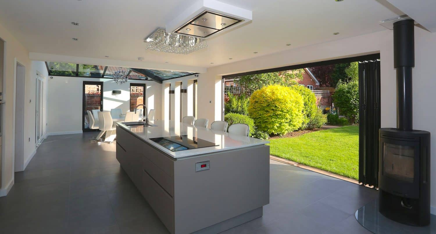 Modern open-plan kitchen with sleek island, glass doors fully opened to a lush green garden, dining area in the background, and a black wood stove on the right. Natural light fills the space.