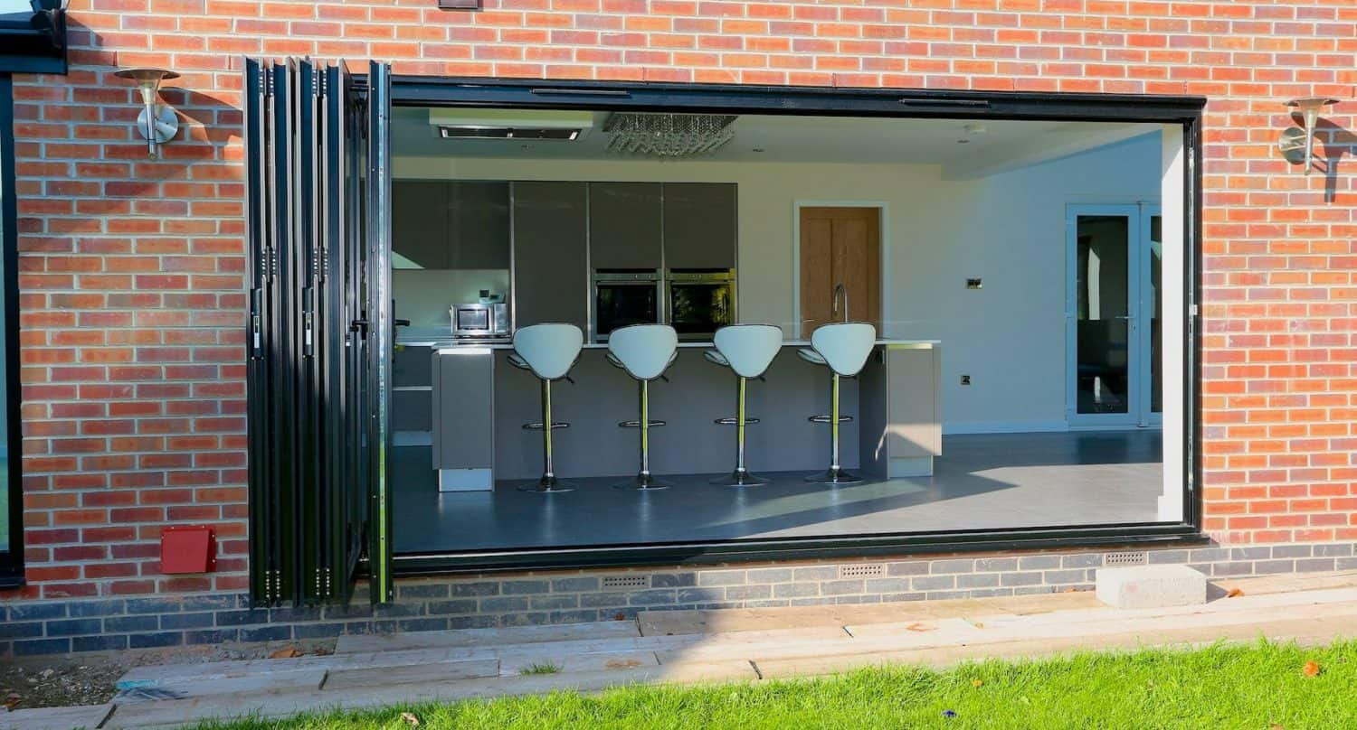 Modern kitchen with a row of five white bar stools at a breakfast bar, viewed through large open folding glass doors set in a brick wall. Green grass and stone path are visible outside.