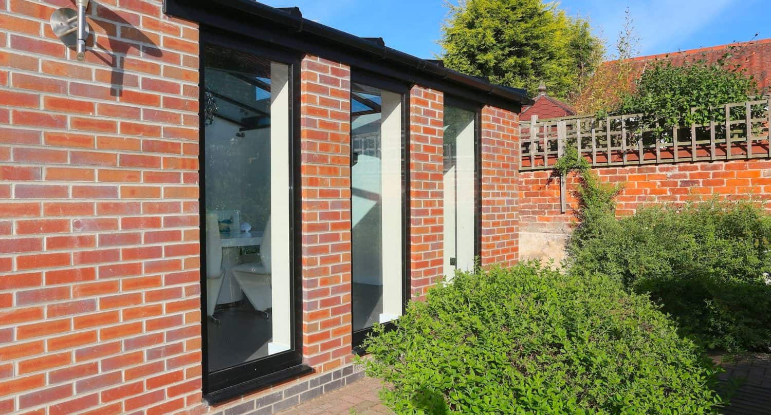 A small brick building with three tall glass windows, surrounded by green bushes and a paved path, with a wooden fence and trees in the background under a blue sky.