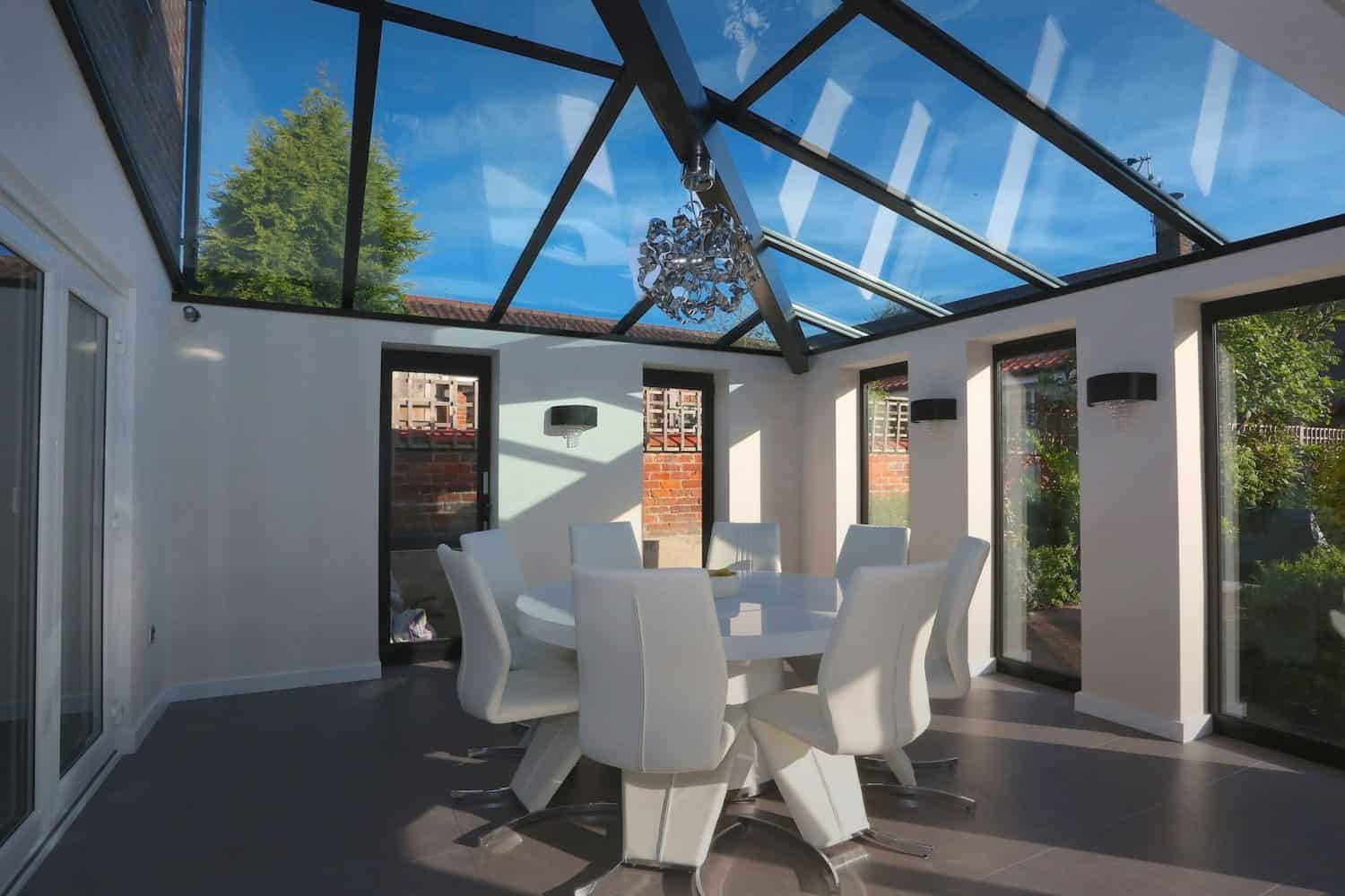 A modern conservatory with a glass roof and walls, featuring a round white dining table surrounded by white chairs, and a chandelier hanging above. Sunlight streams in, highlighting the bright, airy space.