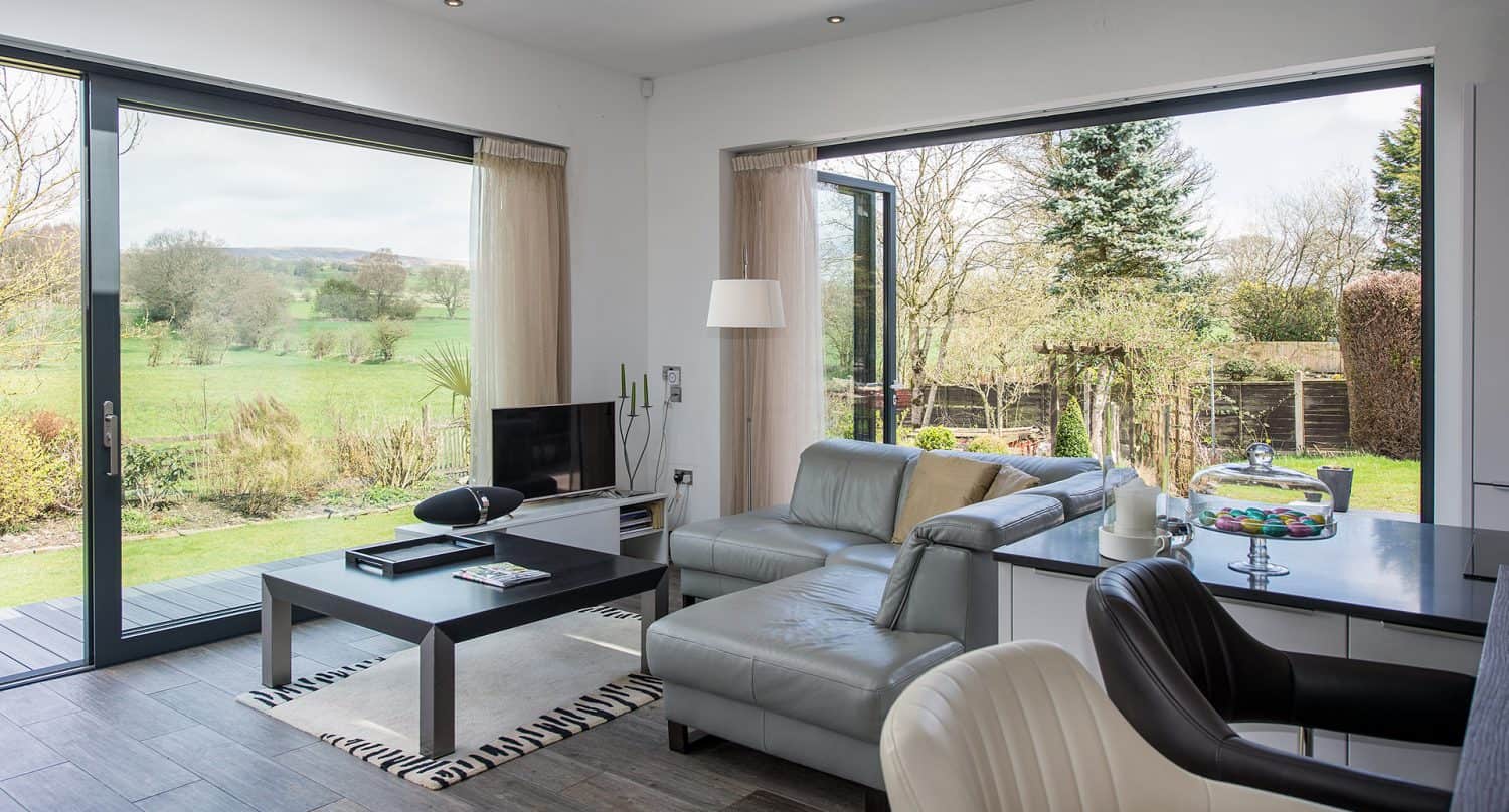 Modern living room with large glass doors and windows overlooking a green garden and fields. The room features a gray sofa, coffee table, and kitchen island, with natural light filling the space.