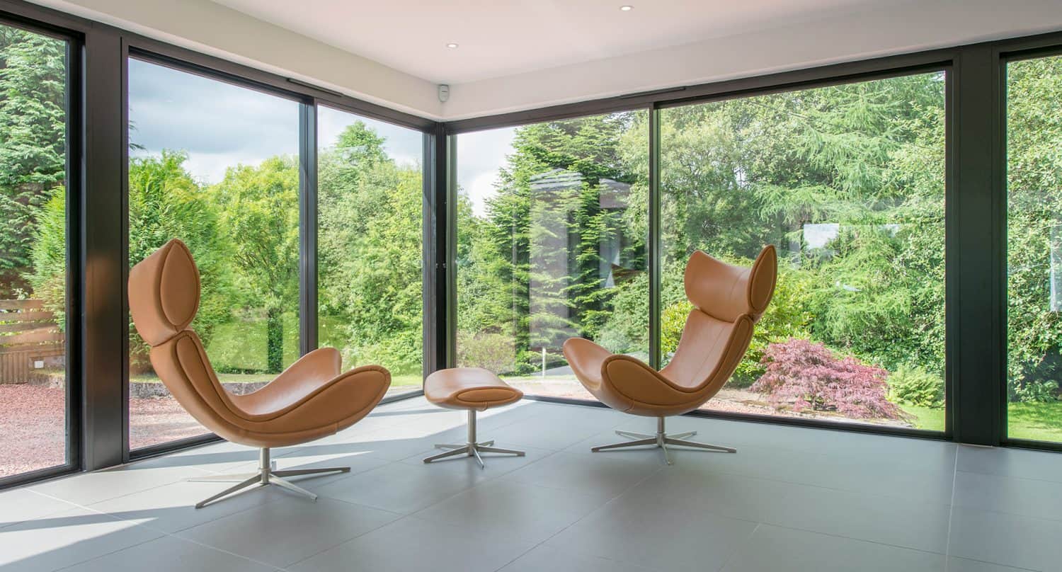 Two modern tan lounge chairs and a matching ottoman sit in a bright, minimalist room with floor-to-ceiling windows, offering views of lush green trees and landscaped gardens outside.
