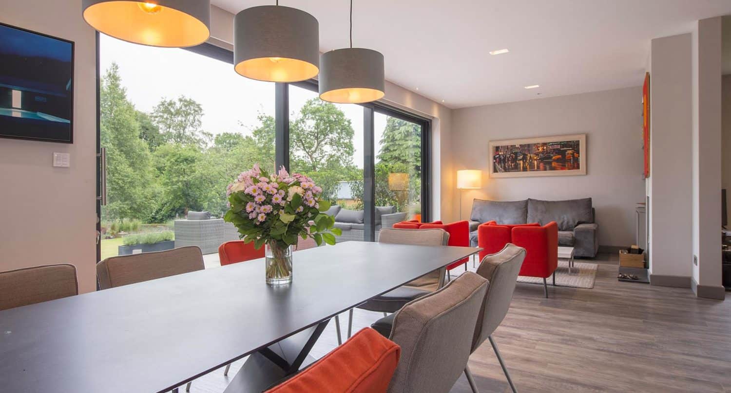 Modern dining and living area with a black table, gray and orange chairs, a vase of flowers, large windows overlooking a green garden, and cozy seating with lamps and artwork on the walls.