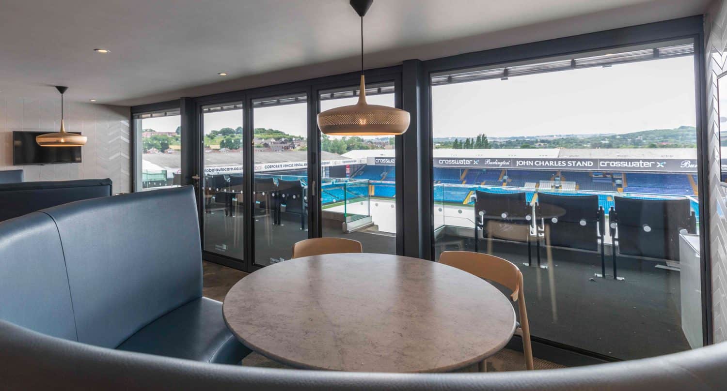 A modern lounge with a round table and booth seating overlooks a stadium through large glass doors, offering a clear view of blue seats and the field outside.
