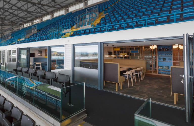 A modern sports stadium suite with rows of blue seats outside and an indoor lounge area featuring tables, chairs, and a bar, separated by glass doors.
