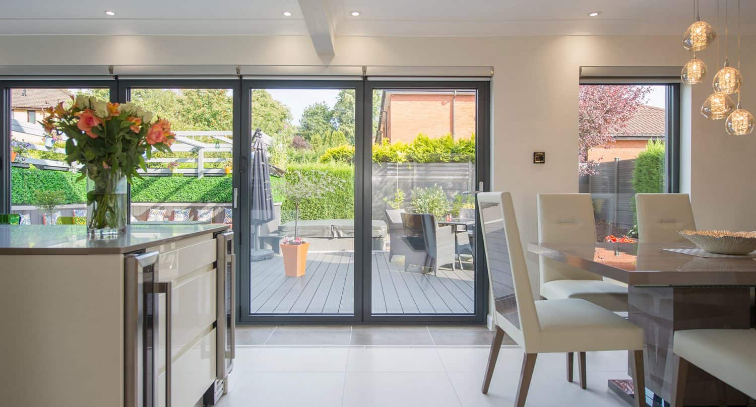 Modern kitchen and dining area with large glass sliding doors leading to a patio with outdoor seating. The space is bright, decorated with flowers, and illuminated by pendant lights.