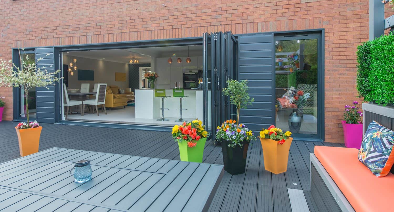 Modern patio with brightly colored flower pots on a gray deck. Large glass doors open to a stylish kitchen and dining area. An orange cushioned bench and green plants add vibrant accents to the space.
