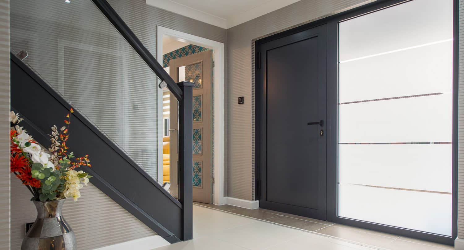 Modern hallway with a black front door, large frosted glass side panel, grey staircase with black railing, and a vase of colorful flowers on the floor. Light walls and bright lighting create an airy atmosphere.
